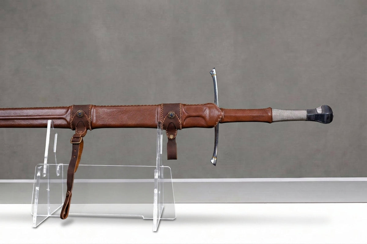 Titanforge Zweihänder : Scheidenmund aus braunem Leder mit Messing-Conchos, genähter Gürtelschlaufe, Parierstange aus Stahl und Griff vor grauem Betonhintergrund
